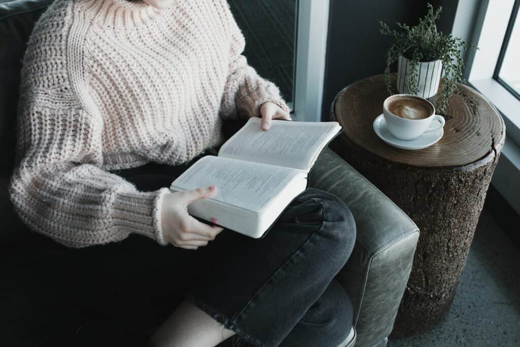 woman in white knit sweater and blue denim jeans sitting on gray couch reading book